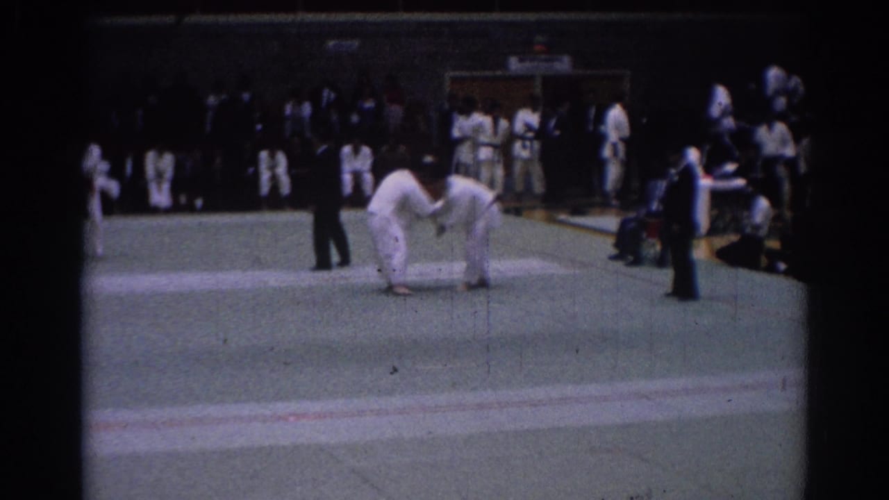 1967 Los Angeles judo tournament frame with two competitors gripping each other on the mat, a referee nearby, and uniformed observers at the edge.