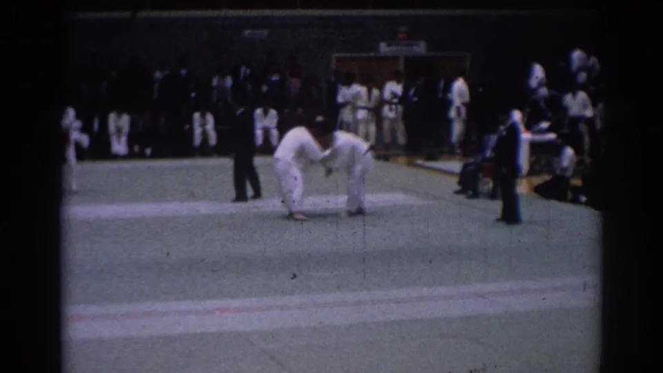 1967 Los Angeles judo tournament frame with two competitors gripping each other on the mat, a referee nearby, and uniformed observers at the edge.