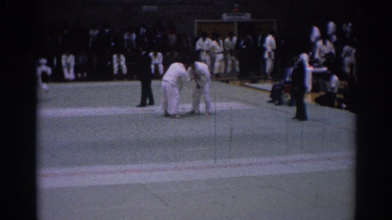 Two judo athletes grapple on an indoor mat while a referee and spectators watch.