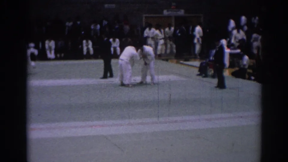 1967 Los Angeles home-movie frame showing two judo competitors in white gi grappling on a green indoor mat with a referee nearby.