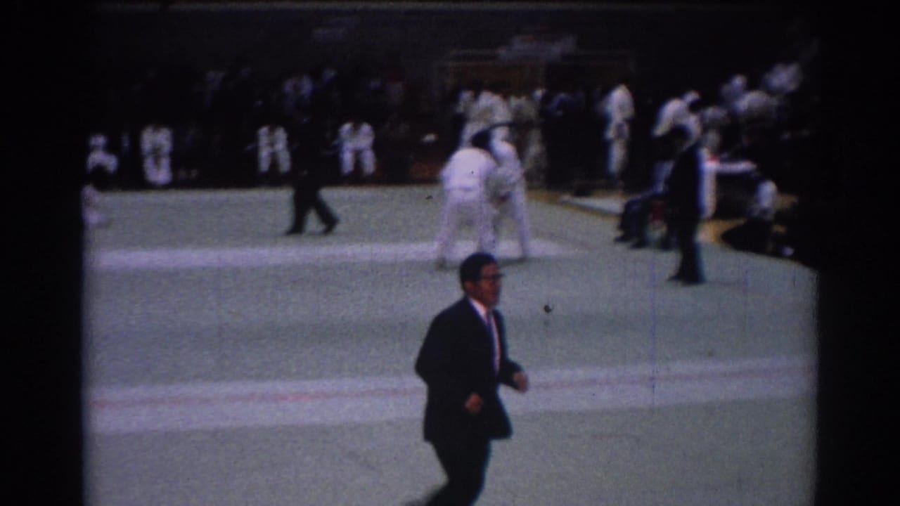 A man in a dark suit walks across the foreground during the 1967 Los Angeles judo competition, with athletes grappling behind him.