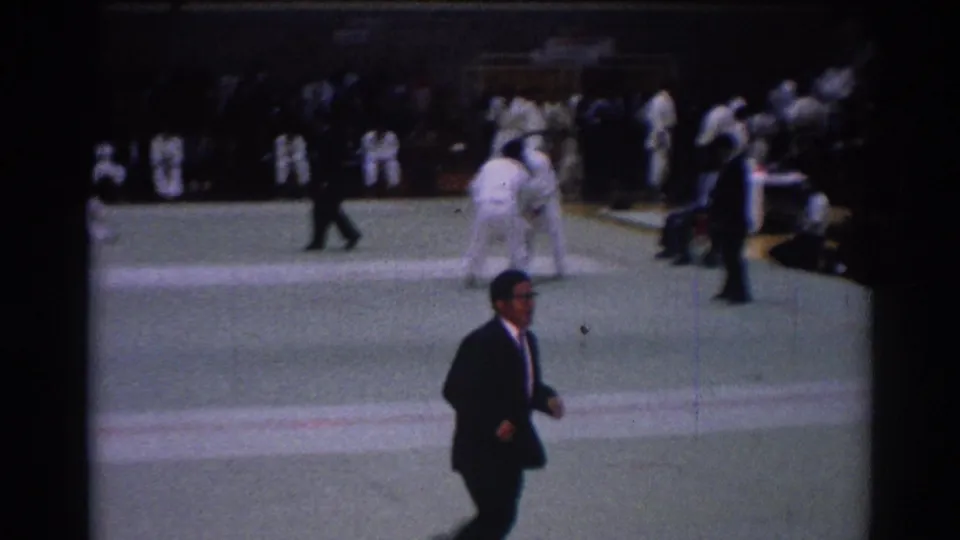 A man in a dark suit walks across the foreground during the 1967 Los Angeles judo competition, with athletes grappling behind him.