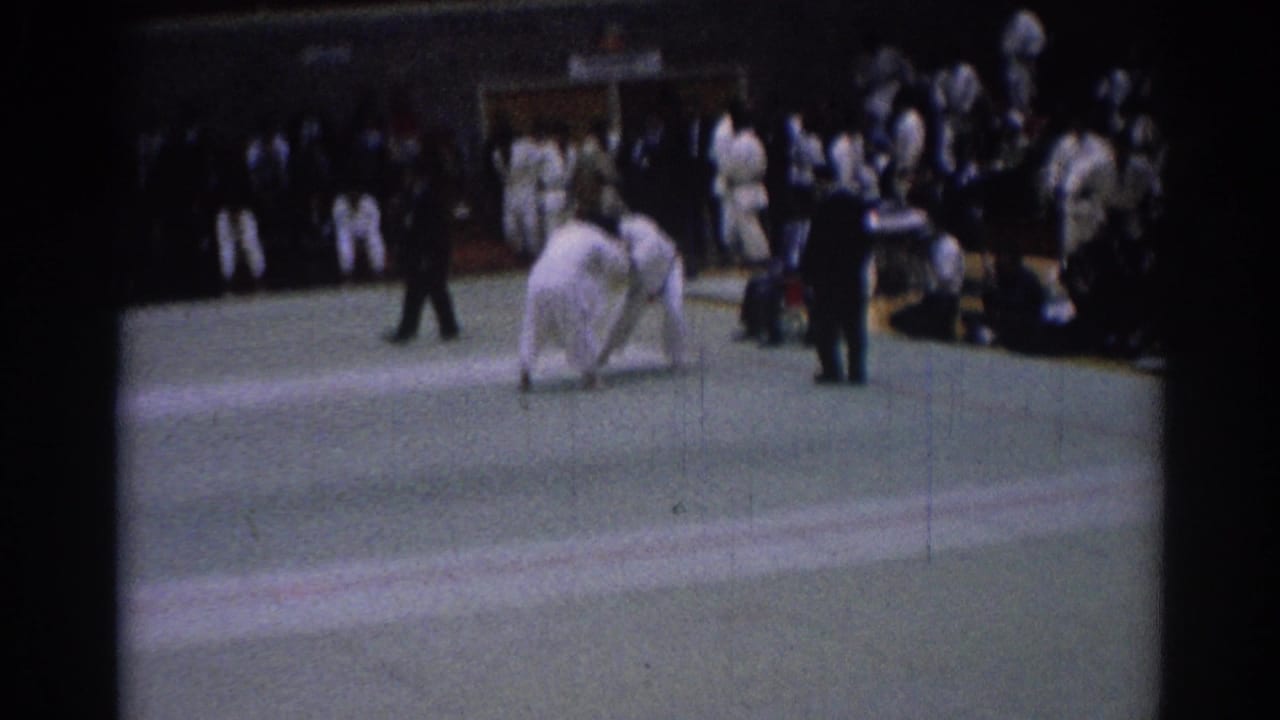 1967 Los Angeles judo competitors in white gi hold a standing grip on the indoor mat as a referee and spectators observe.