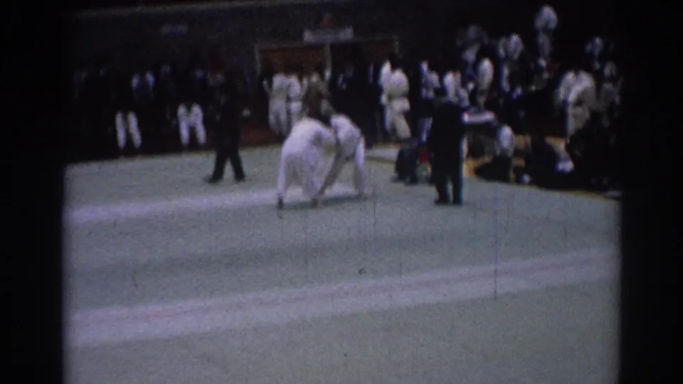 1967 Los Angeles judo competitors in white gi hold a standing grip on the indoor mat as a referee and spectators observe.