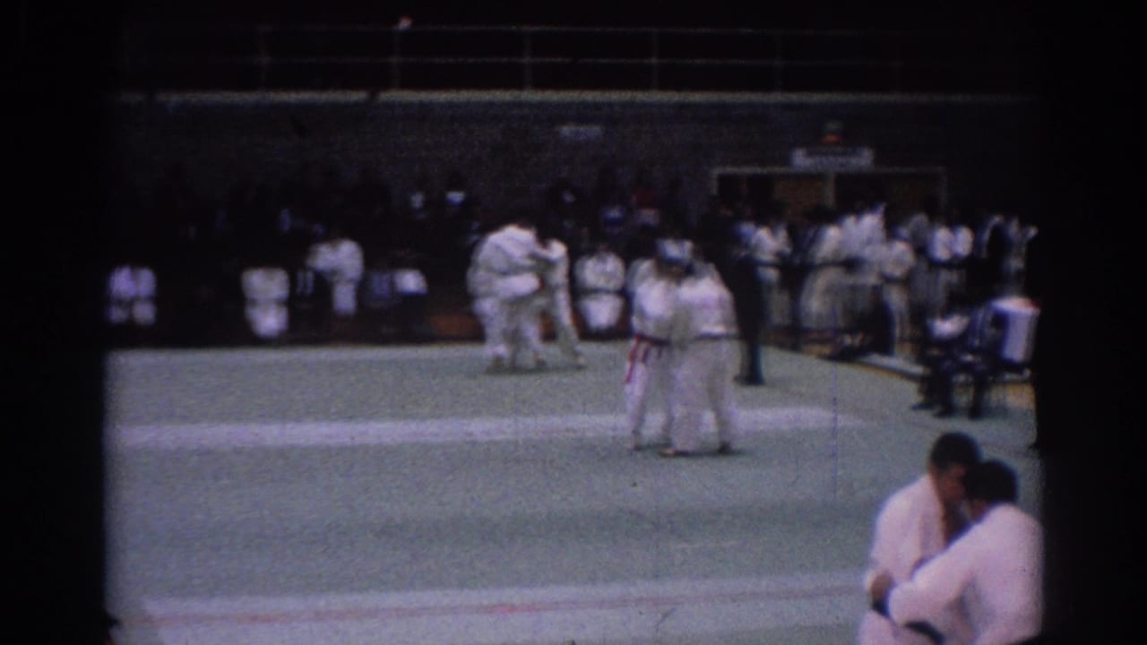 Wider 1967 Los Angeles gym view with judo competitors, a referee, spectators, and two white-gi athletes grappling on the mat.