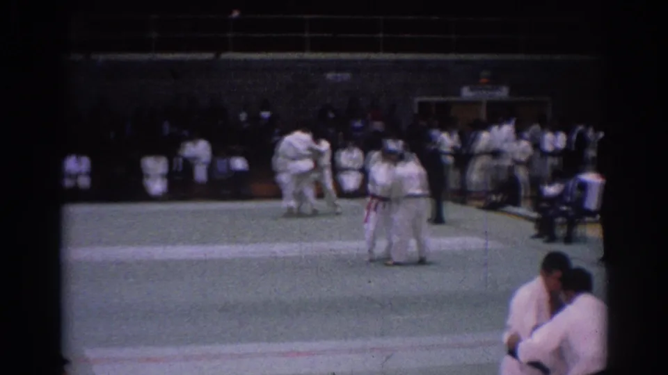 Wider 1967 Los Angeles gym view with judo competitors, a referee, spectators, and two white-gi athletes grappling on the mat.