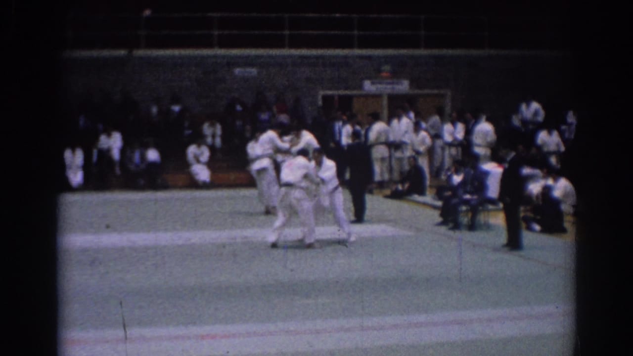 Two martial arts competitors in white gi grapple during a 1967 Los Angeles indoor tournament scene.