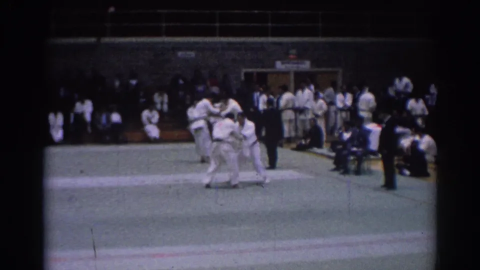 Two martial arts competitors in white gi grapple during a 1967 Los Angeles indoor tournament scene.