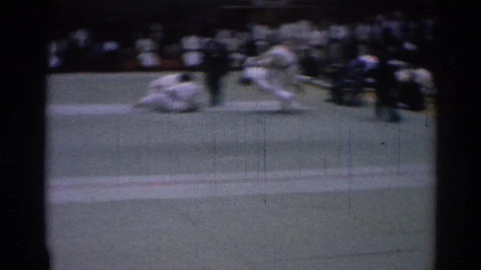 A blurred 8mm frame shows one martial arts competitor on the mat while another moves away during the bout.