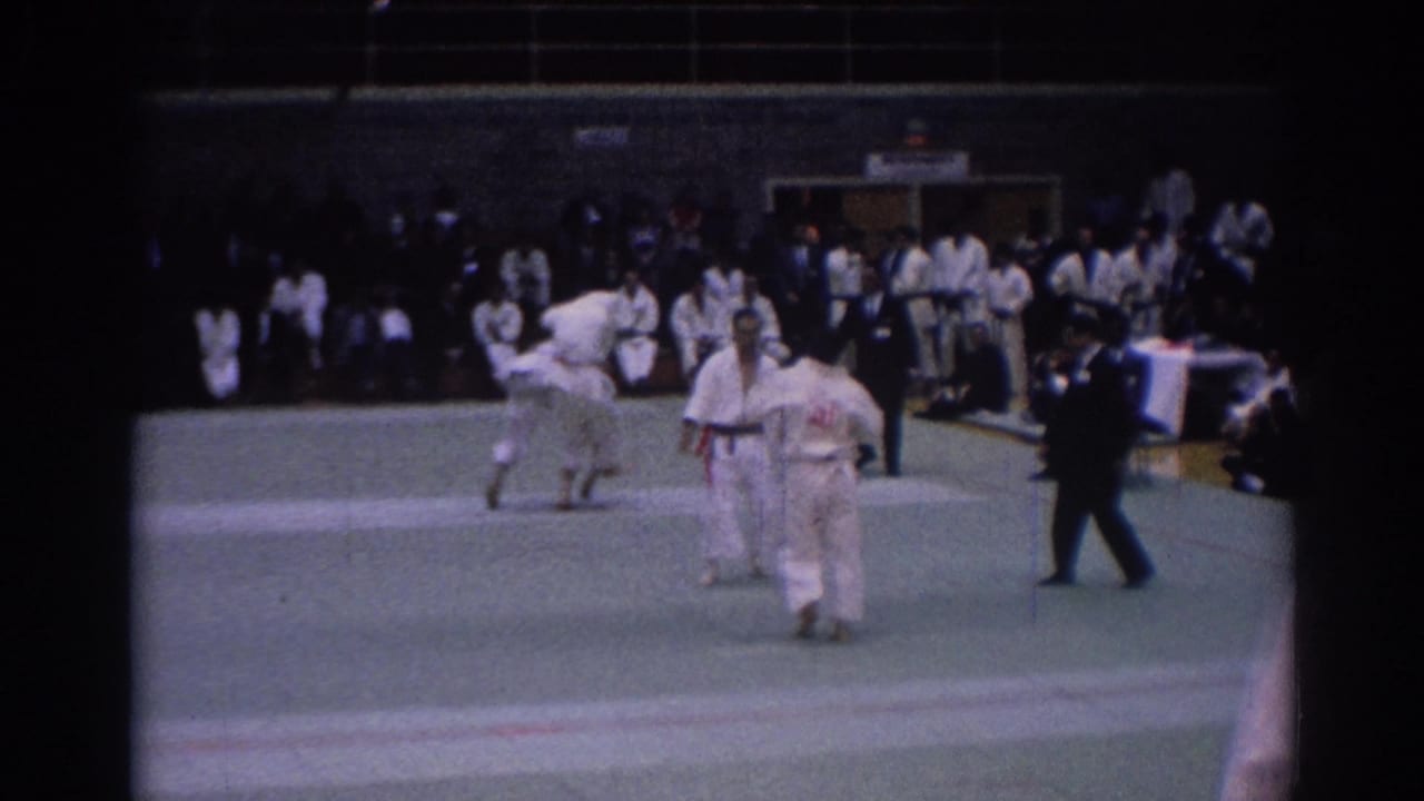 The 1967 Los Angeles martial arts match pauses with competitors and referee on the indoor mat before seated spectators.
