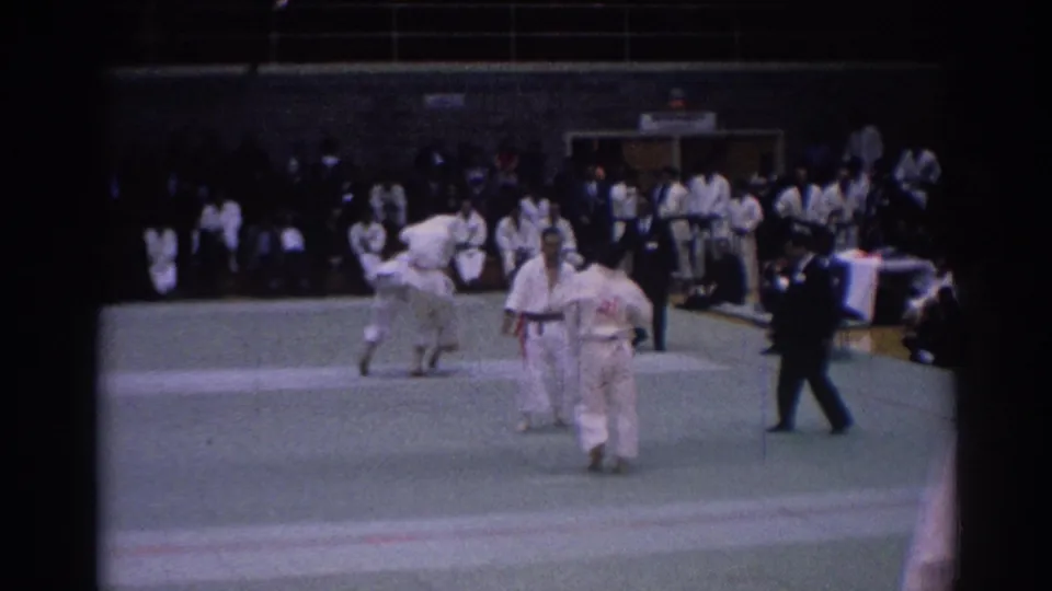 The 1967 Los Angeles martial arts match pauses with competitors and referee on the indoor mat before seated spectators.