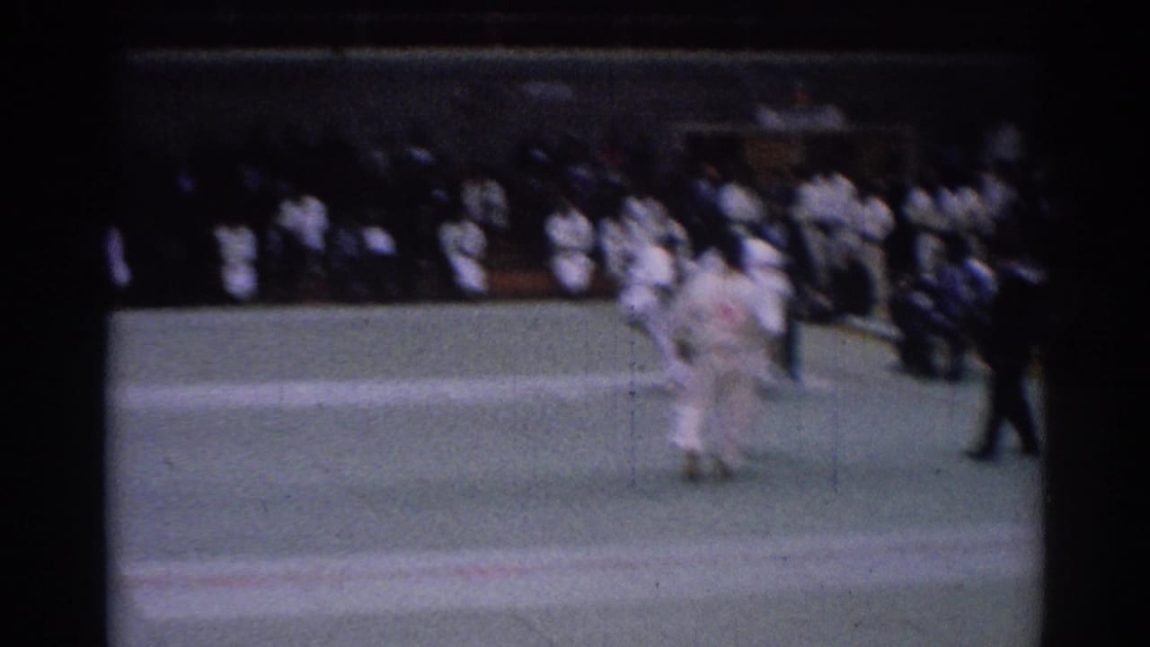 Two karate competitors in white gi grapple on a green mat as a referee and spectators watch.