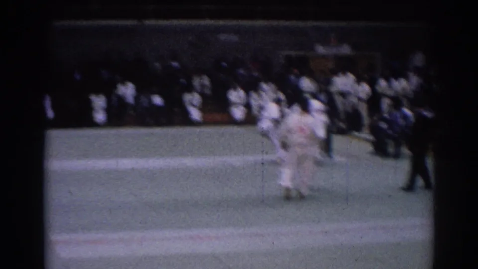 1967 Los Angeles home-movie frame showing a karate match on a green mat, with uniformed spectators seated along the far side.