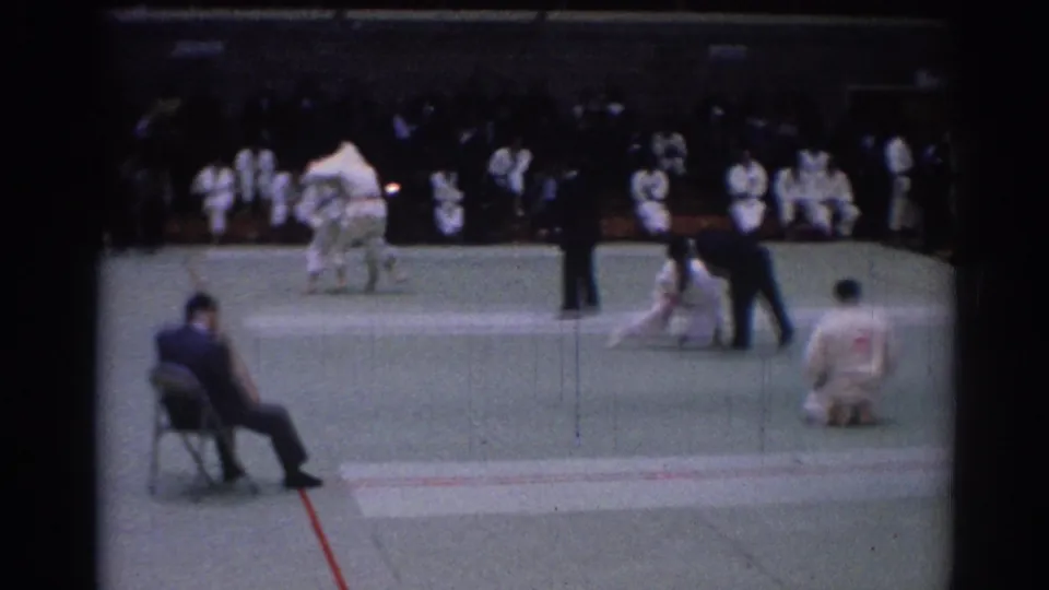 A 1967 Los Angeles karate tournament scene shows competitors on the mat, a seated judge, referees, and waiting participants in white gi.