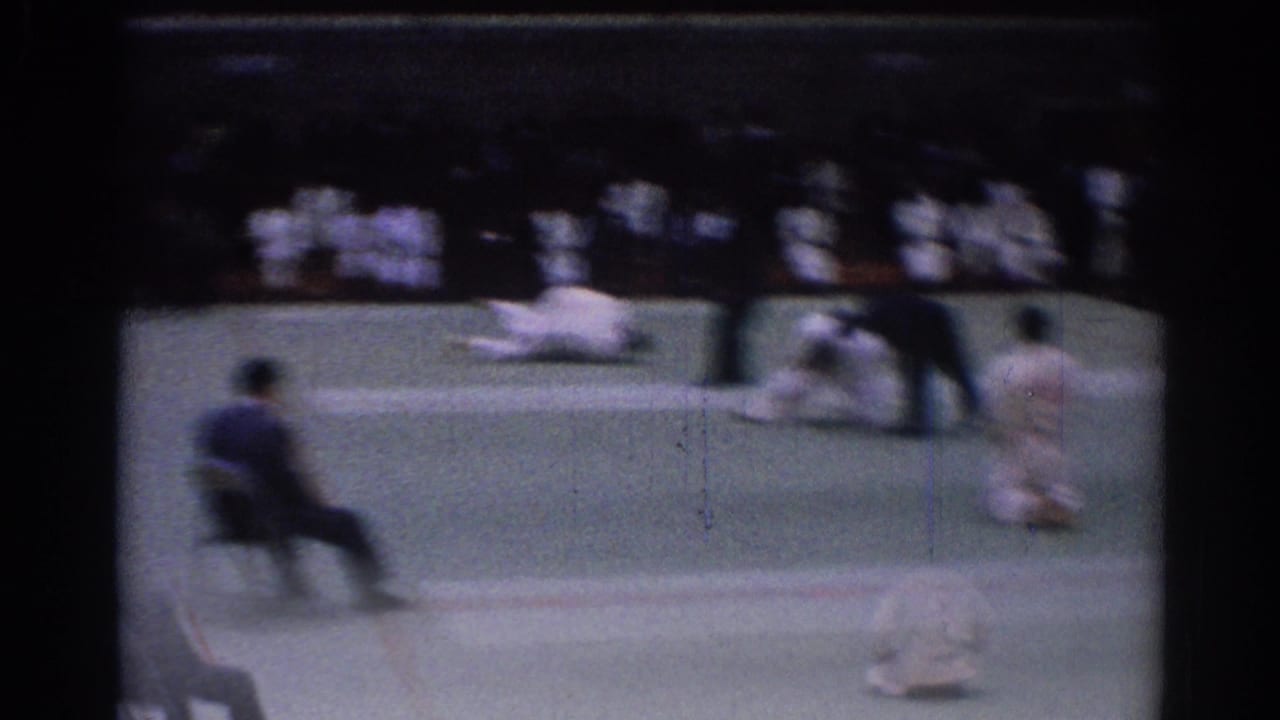 Motion-blurred 1967 Los Angeles karate tournament frame with a competitor low on the mat, officials nearby, and uniformed spectators behind.