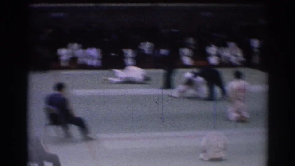 Motion-blurred 1967 Los Angeles karate tournament frame with a competitor low on the mat, officials nearby, and uniformed spectators behind.