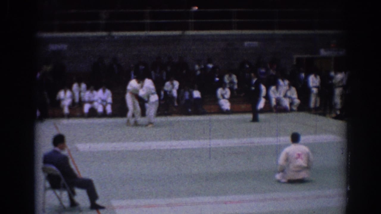 1967 Los Angeles home-movie frame showing two karate competitors reset on the mat, with referees, a seated judge, and spectators in white gi.