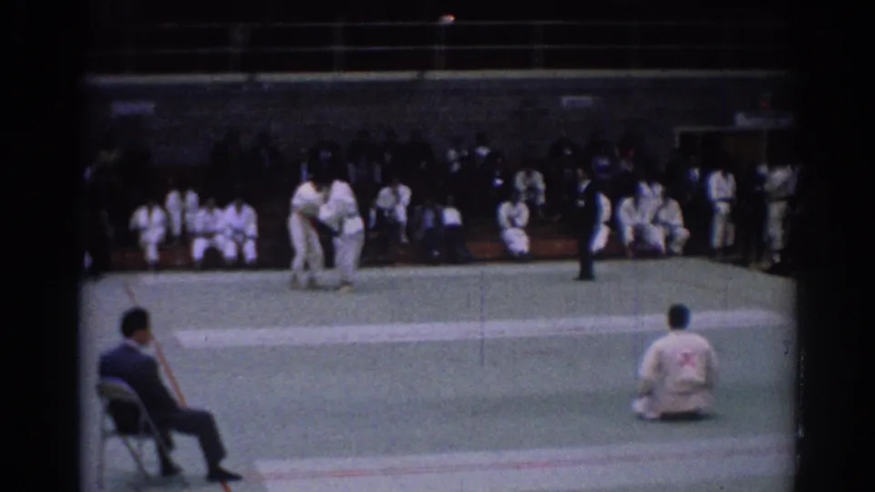 1967 Los Angeles home-movie frame showing two karate competitors reset on the mat, with referees, a seated judge, and spectators in white gi.
