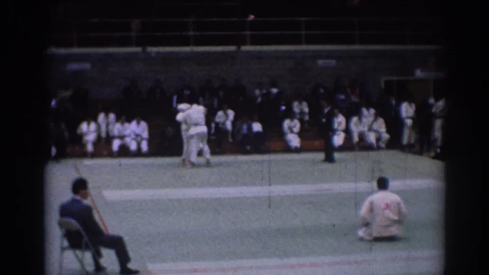 1967 Los Angeles 8mm frame showing two judoka on a green mat with a referee nearby and spectators seated along the wall.