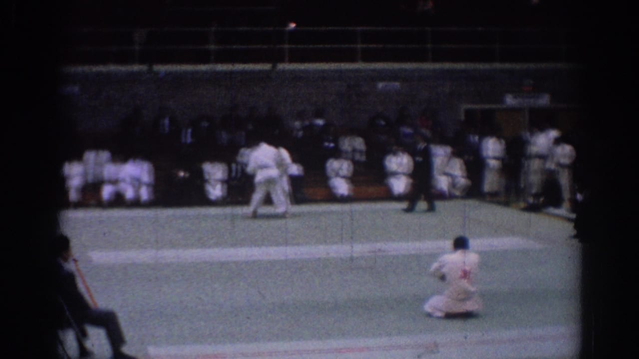1967 Los Angeles judo competition frame with two athletes in white gi, a referee, and seated spectators in an indoor venue.