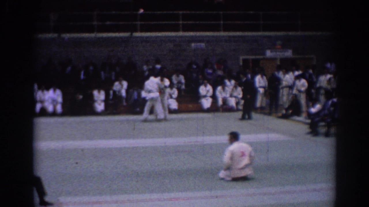 1967 Los Angeles 8mm still with a seated judoka in the foreground, competitors on the mat, and spectators along the indoor wall.