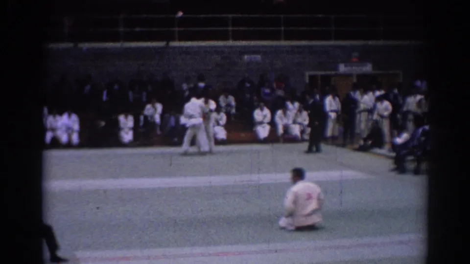 1967 Los Angeles 8mm still with a seated judoka in the foreground, competitors on the mat, and spectators along the indoor wall.