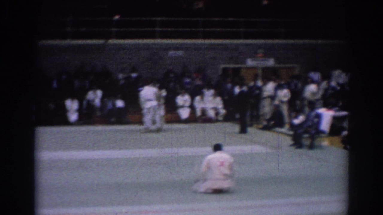 1967 Los Angeles competition frame showing judoka on a green mat, a dark-suited referee, and spectators seated around the venue.