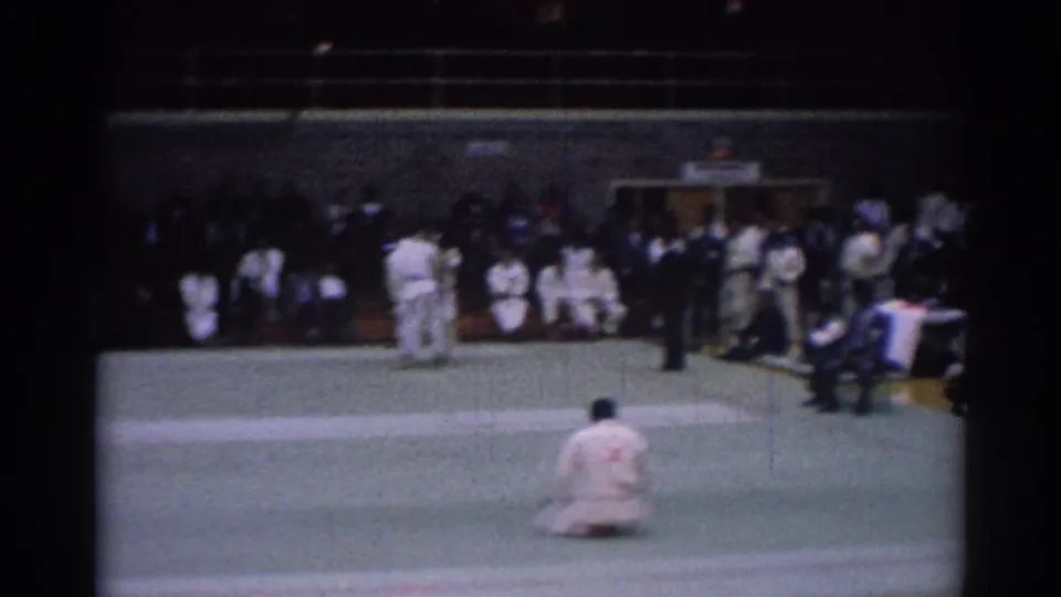 1967 Los Angeles competition frame showing judoka on a green mat, a dark-suited referee, and spectators seated around the venue.