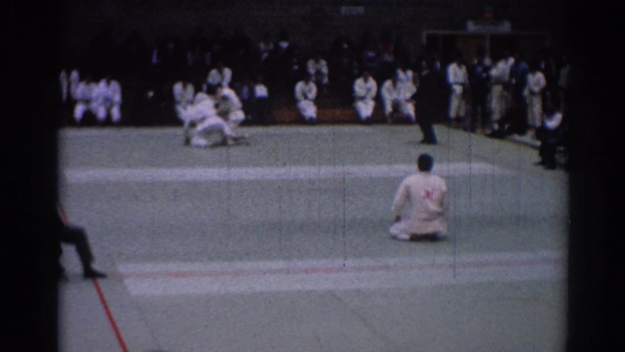 Two karate competitors grapple on a green mat as a seated participant and spectators watch.