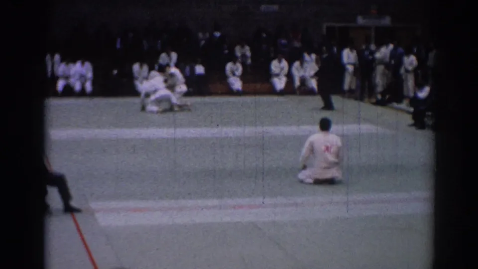 1967 Los Angeles home-movie frame of karate competitors grappling on a green mat with seated participants and spectators in a gym.