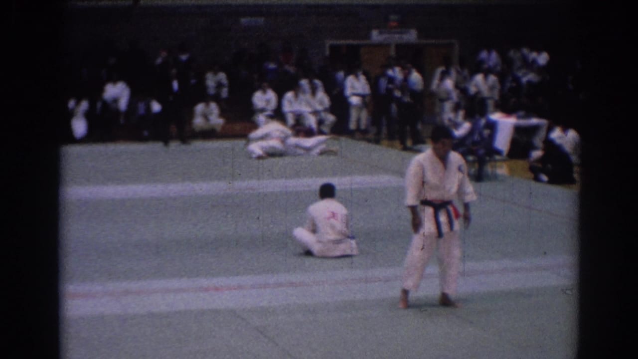 1967 Los Angeles karate match frame with a dark-belt referee standing in the foreground as competitors complete a takedown behind him.