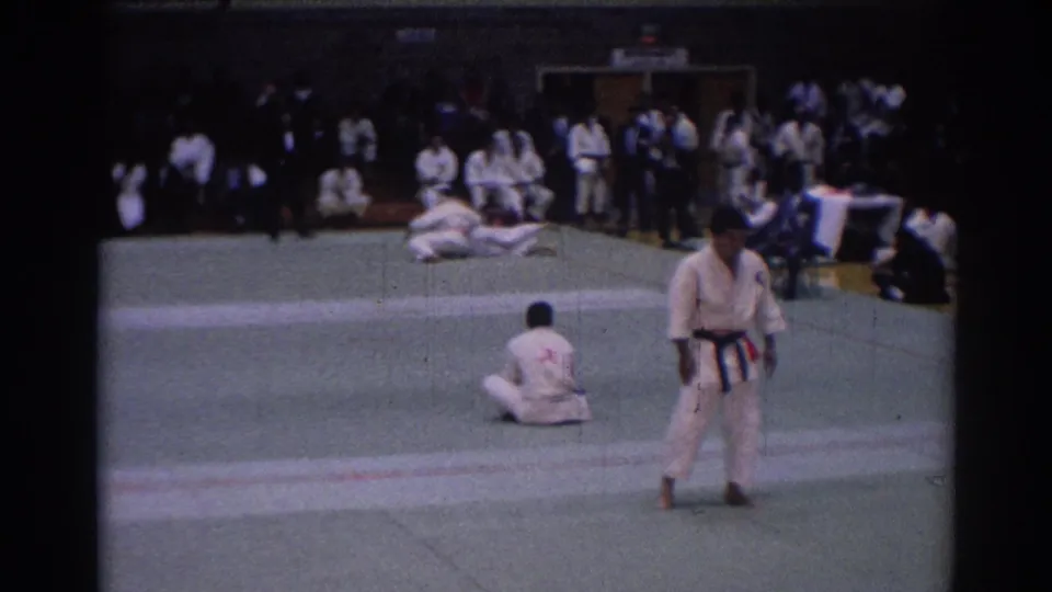 1967 Los Angeles karate match frame with a dark-belt referee standing in the foreground as competitors complete a takedown behind him.