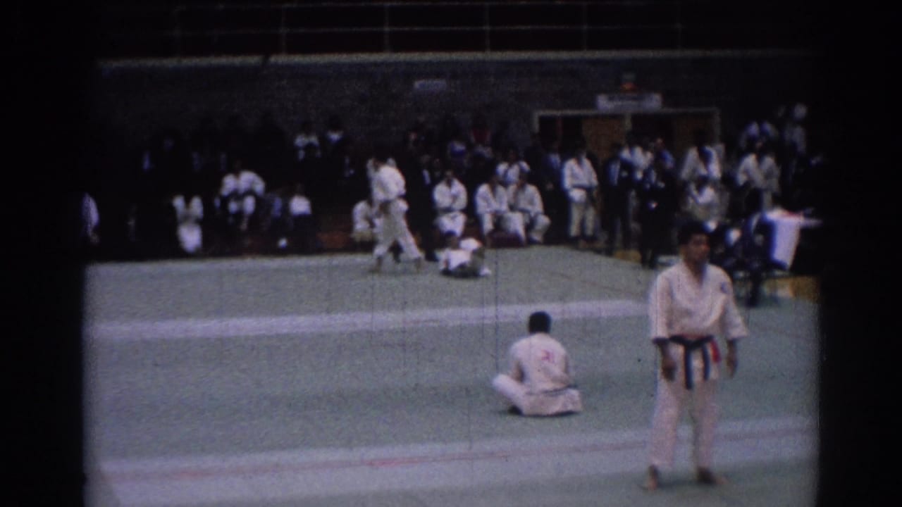 Los Angeles 1967 karate competition frame showing a referee on the mat with one competitor down and seated participants along the gym wall.