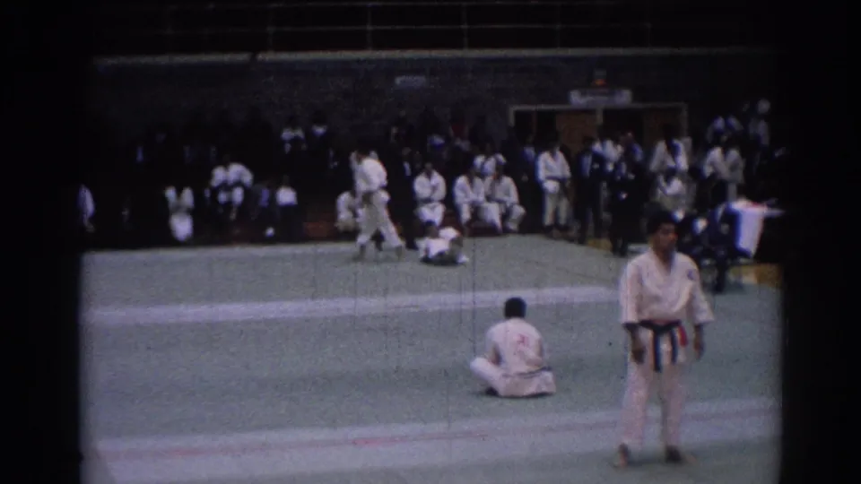 Los Angeles 1967 karate competition frame showing a referee on the mat with one competitor down and seated participants along the gym wall.