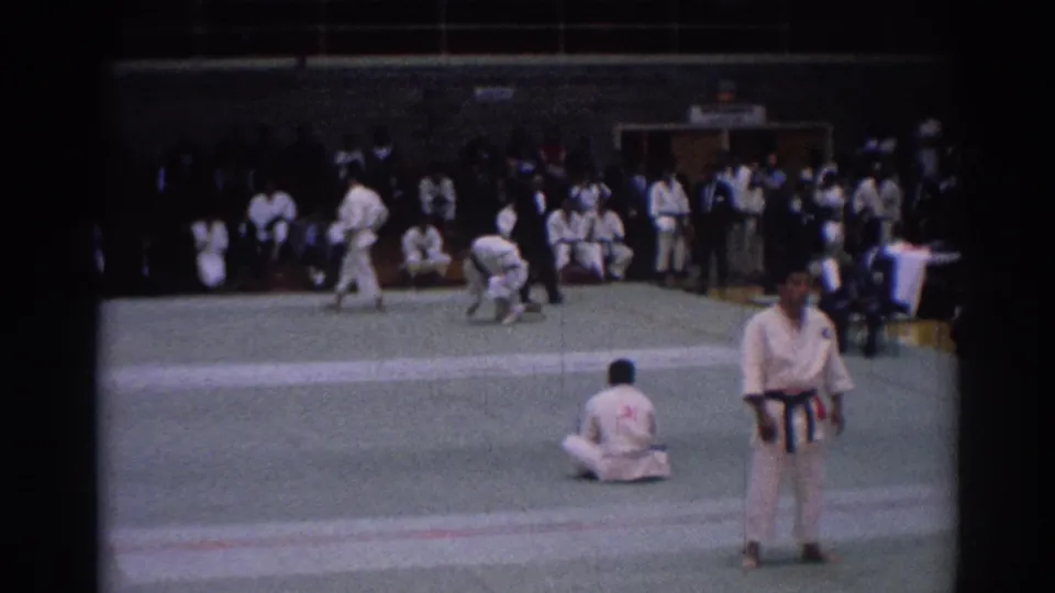 1967 Los Angeles 8mm frame of karate competitors on an indoor green mat with seated spectators and an official nearby.