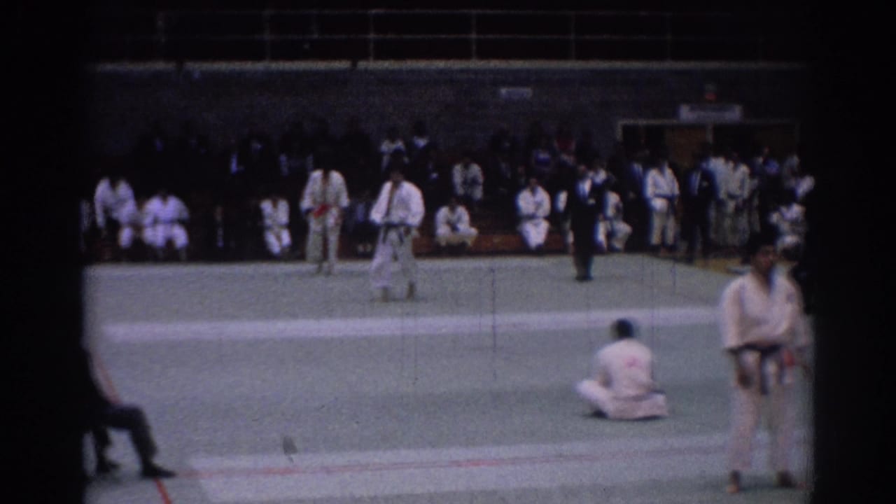 1967 Los Angeles tournament frame showing gi-clad karate fighters on a green indoor mat with a dark-suited official and audience.