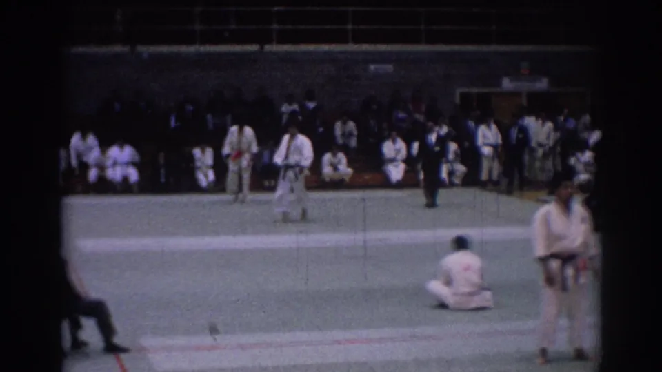 1967 Los Angeles tournament frame showing gi-clad karate fighters on a green indoor mat with a dark-suited official and audience.