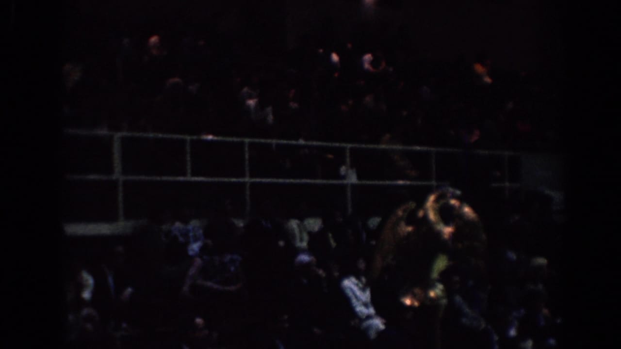 Dim 1967 Los Angeles tournament frame showing spectators in bleachers behind railings, filmed from a handheld 8mm viewpoint.