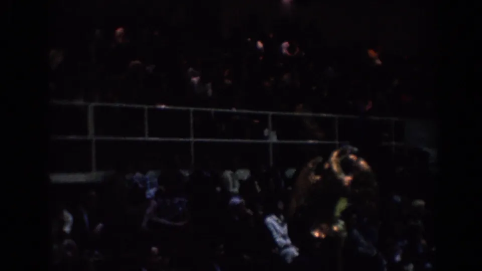 Dim 1967 Los Angeles tournament frame showing spectators in bleachers behind railings, filmed from a handheld 8mm viewpoint.
