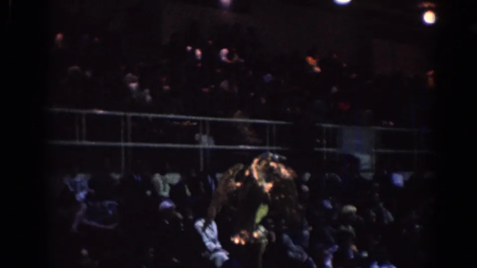 1967 Los Angeles home-movie frame showing spectators in indoor bleachers and a bright gold eagle-like symbol near the competition area.