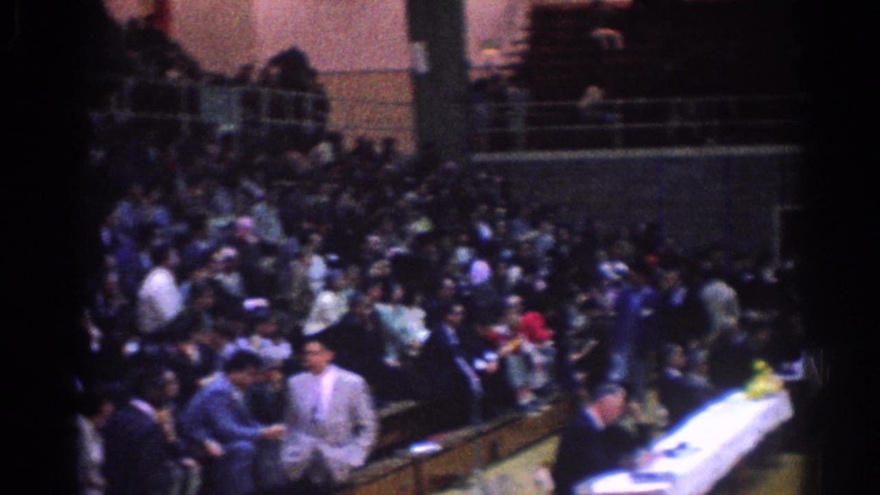 Home-movie view of a 1967 Los Angeles indoor martial arts event, with spectators, suited men, and a white-covered table near the mat.