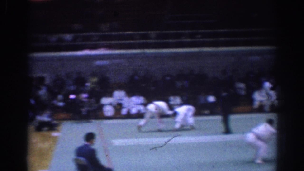 1967 Los Angeles martial arts match frame showing competitors in white uniforms on a green mat with spectators seated beyond them.