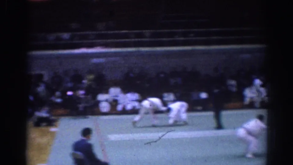 1967 Los Angeles martial arts match frame showing competitors in white uniforms on a green mat with spectators seated beyond them.