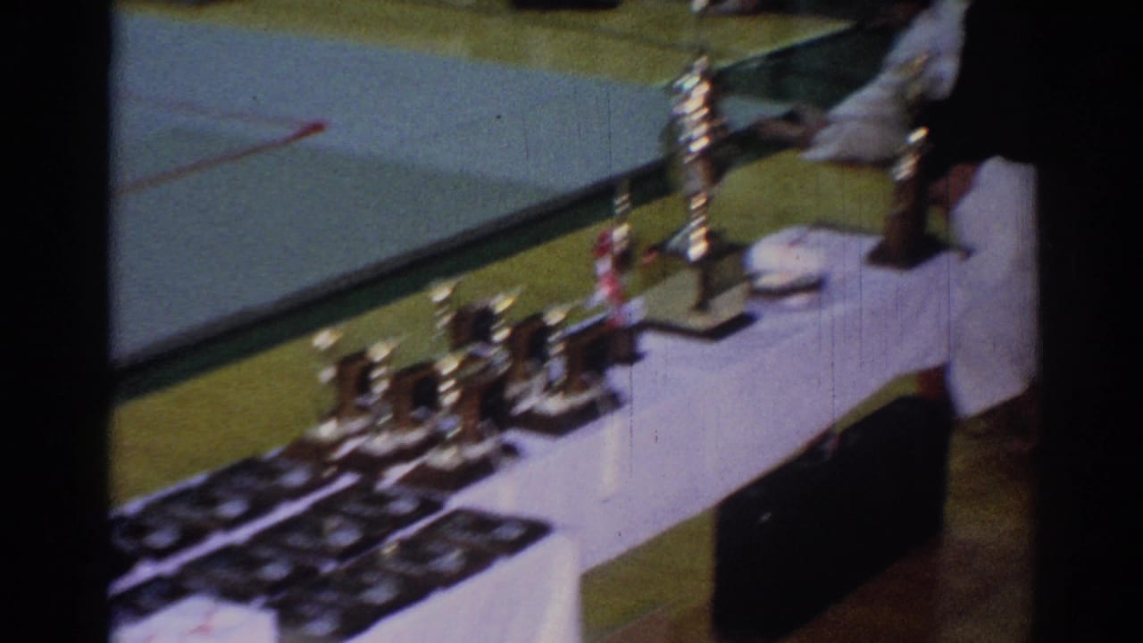 1967 Los Angeles home-movie frame showing a white-draped awards table with trophies, plaques, and a person in a gi nearby.