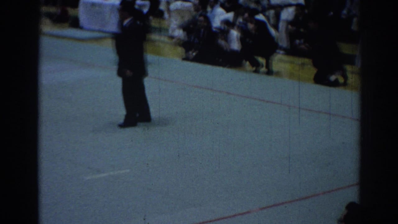 A man in a dark suit stands on the green competition floor while spectators sit along the edge of the Los Angeles venue.