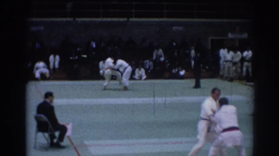 1967 Los Angeles judo match on 8mm film, with athletes in white gi grappling on a green mat before spectators.