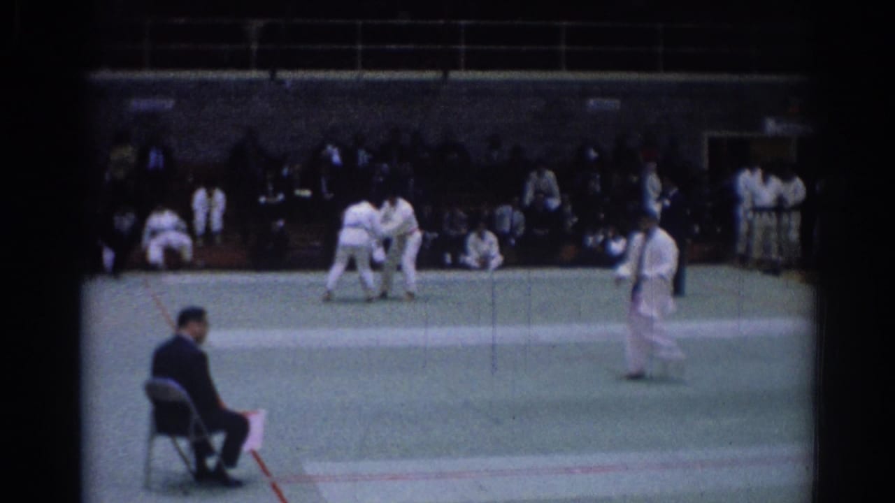 Indoor 1967 Los Angeles judo competition scene with grappling athletes, a standing official, and seated spectators.