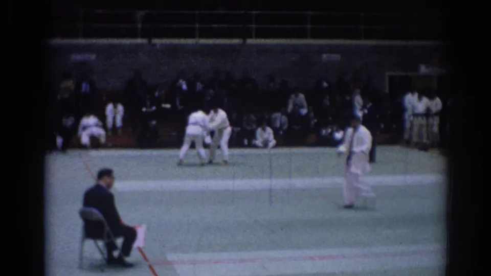 Indoor 1967 Los Angeles judo competition scene with grappling athletes, a standing official, and seated spectators.
