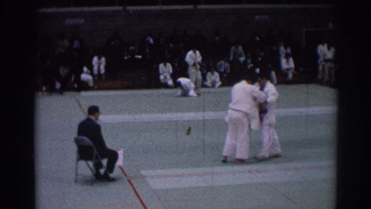 1967 Los Angeles 8mm still showing two judo athletes in white gi grappling before bleachers of spectators.