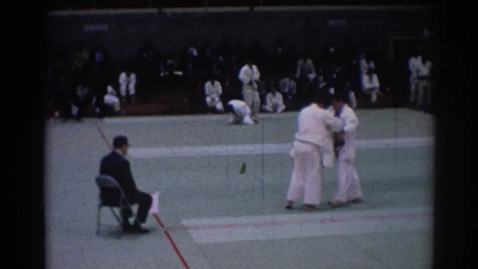 1967 Los Angeles 8mm still showing two judo athletes in white gi grappling before bleachers of spectators.